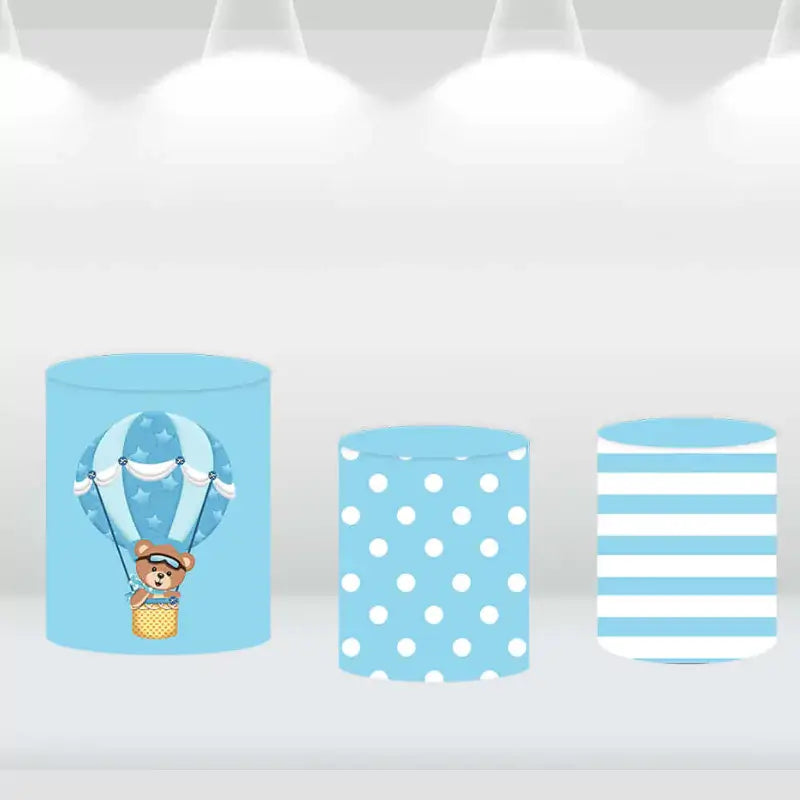 Three cylindrical covers featuring blue and white polka dots, stripes, and hot air balloon patterns, designed to complement a teddy bear backdrop.