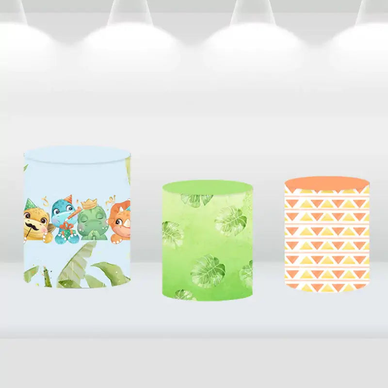 Set of three cylinder covers featuring cute dino designs, tropical leaves, and geometric patterns, matching the dinosaur-themed party backdrop.