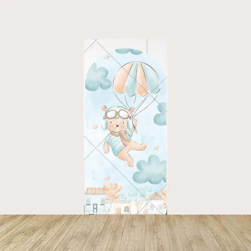 Arch panel featuring a teddy bear in aviator gear holding parachutes, floating through watercolor clouds above a row of pastel houses.
