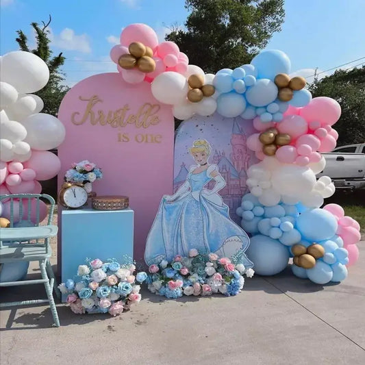 Cinderella first birthday party backdrop with pink and blue arches, balloon garlands, name sign “Kristelle is one,” castle illustration, and flower decor.