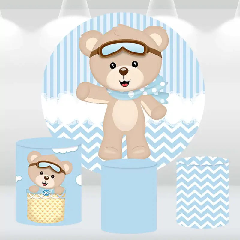 Aviator teddy bear-themed party decor set with a round backdrop featuring a teddy bear in aviator gear and matching blue and white cylinder covers.