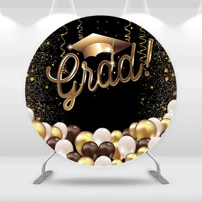 Elegant graduation-themed backdrop with a black and gold design, featuring a large 'Grad!' sign, a gold graduation cap, and a balloon arrangement in gold, black, and white.