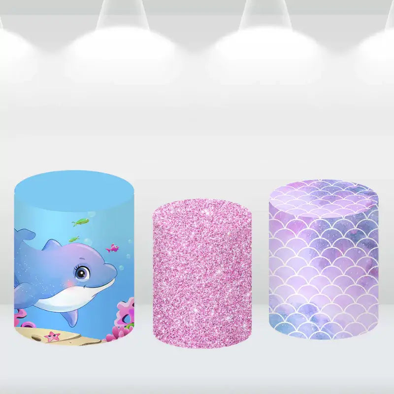 Ocean-themed cylinder covers with designs of a dolphin, pink glitter, and purple mermaid scales, complementing the mermaid party backdrop.