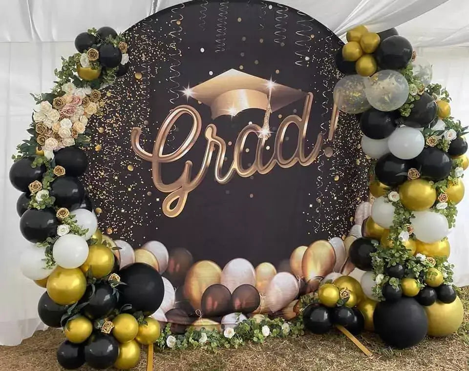 Sophisticated graduation backdrop with black and gold colors, including a 'Grad!' sign, a gold graduation cap, and a cluster of balloons in matching colors.