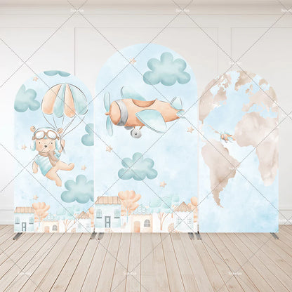 Three-panel aviator-themed arch backdrop featuring a teddy bear with parachute, flying airplane, world map, clouds, and pastel village scene.