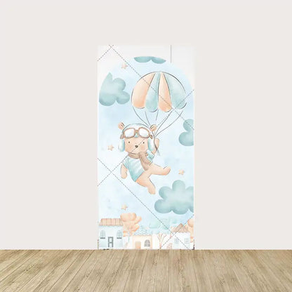 Arch panel featuring a teddy bear in aviator gear holding parachutes, floating through watercolor clouds above a row of pastel houses.