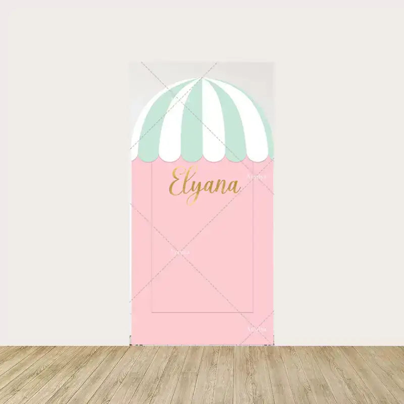 Center panel with a pastel pink backdrop, topped with a striped awning and personalized with "Elyana" in gold script, creating a sweet shop entrance feel.