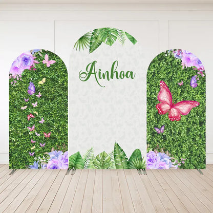Three-panel personalized butterfly garden arch backdrop with hedge greenery, tropical leaves, purple and pink butterflies, and the name “Ainhoa.”