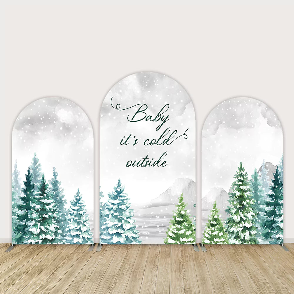 Three-piece arched backdrop set featuring snowy pine trees, soft mountains, and the phrase 'Baby It's Cold Outside' in elegant script, with falling snow creating a winter forest theme.