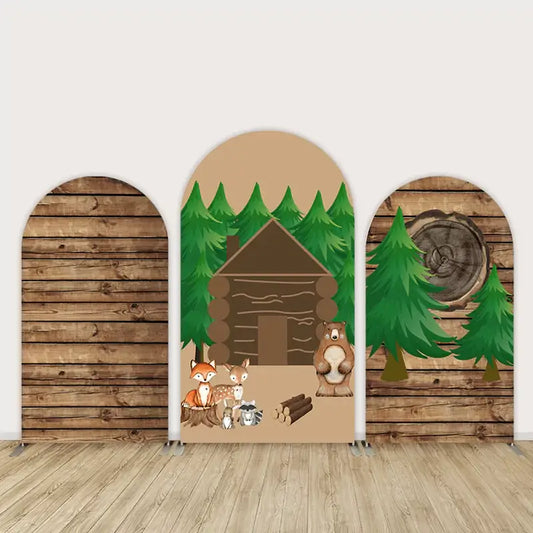Three-panel woodland party arch backdrop featuring a brown log cabin, evergreen trees, and cute animals like a fox, deer, raccoon, and bear with rustic wood textures.